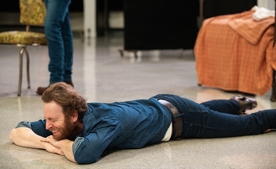 Nick lays on the floor on his stomach, resting his chin on his folded arms. He laughs with his eyes closed.