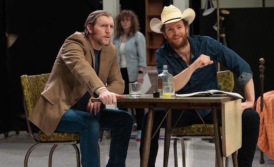 Nick rehearsing with ensemble member Cliff Chamberlain. Cliff is a light-skinned man with a goatee. The two talk at a table as Caroline looks on from the background.