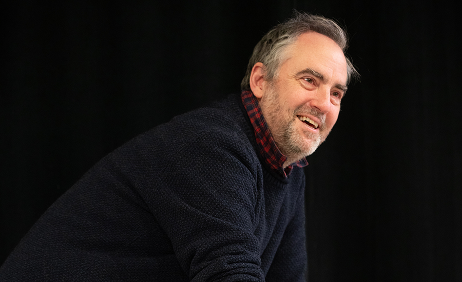 A waist-up photo of Jeremy with a dark background. He is sitting and leaning forward, smiling as he talks.