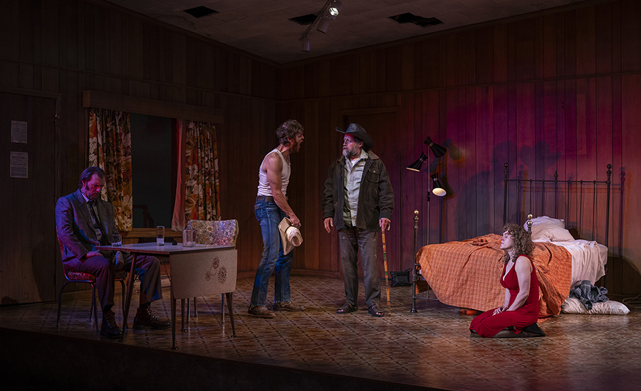 A wide shot of all four characters in the motel room. The Old Man, a light-skinned man with a beard and a cane, stands center stage with Eddie. May kneels on the floor, and Martin sits at the table.