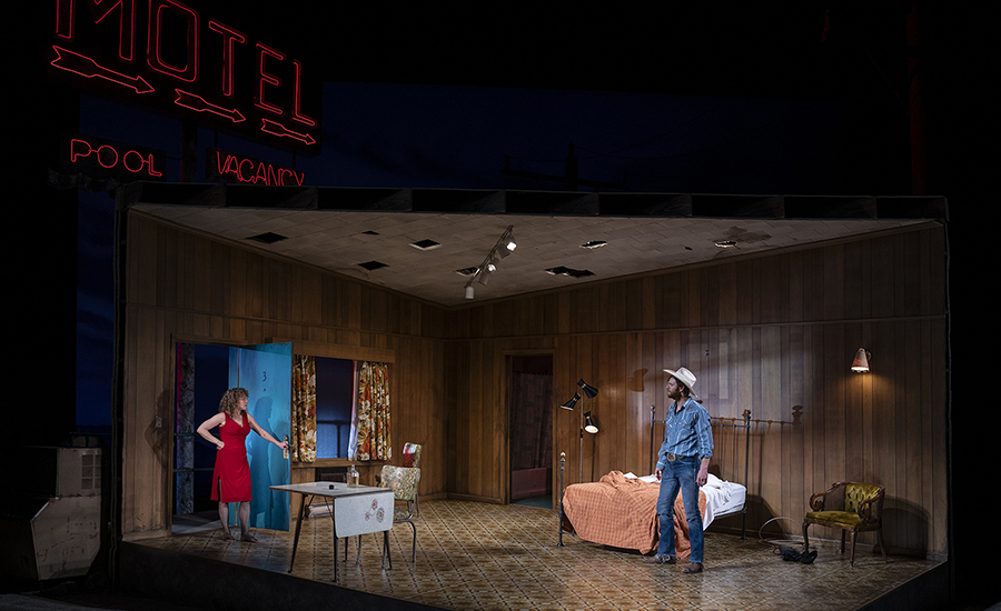A wide shot of Eddie and May, with a red neon motel sign visible in the background. May stands in the doorframe wearing a red dress, and Eddie looks at her from across the room.