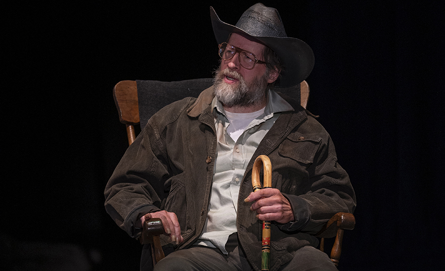 The Old Man sits in his rocking chair, holding his cane. He wears a cowboy hat and is speaking to another character out of frame.
