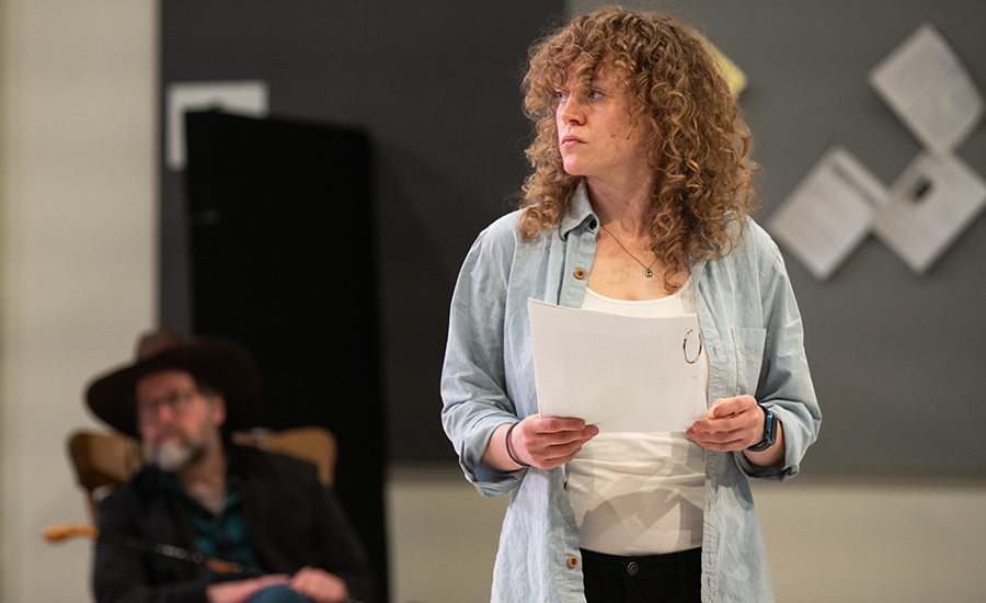 Caroline in rehearsal, looking to the left and holding a script. She wears a blue button down over a white shirt.