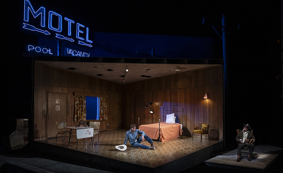  A wide shot of the stage, with the neon motel sign lit up blue. Eddie sits on the floor center stage talks to The Old Man, who sits in his rocking chair on the far side of stage right.