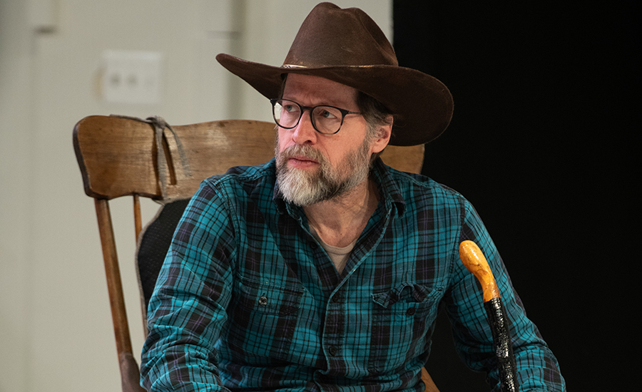 Tim in rehearsal, looking to the left. He sits in a rocking chair, wearing a blue flannel and a cowboy hat.