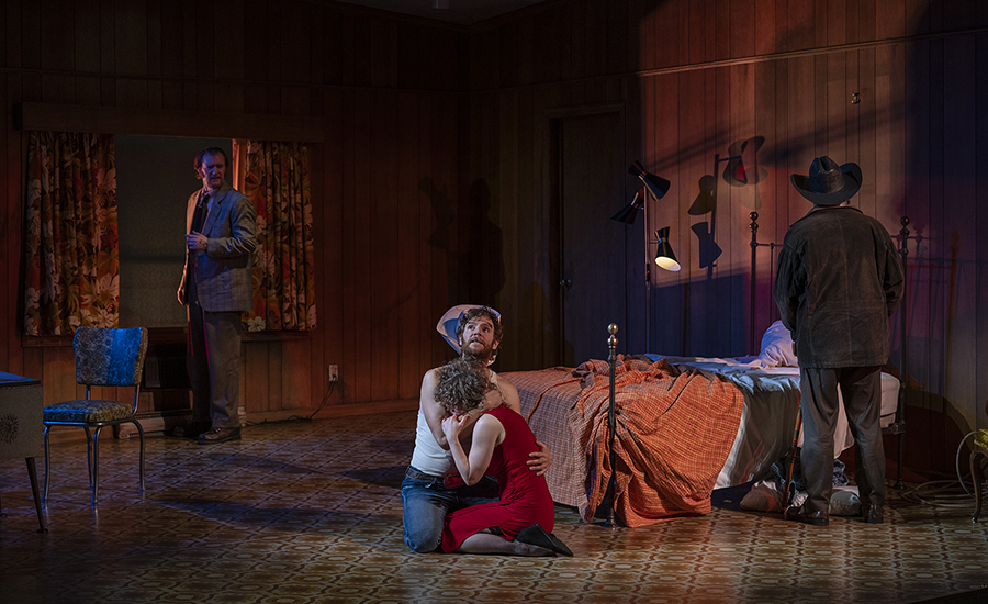 A wide shot of Eddie and May embracing on the floor of the motel room. Martin, a light-skinned man wearing a grey suit, stands at the window. The Old Man stands facing away from the audience, staring at the wall.