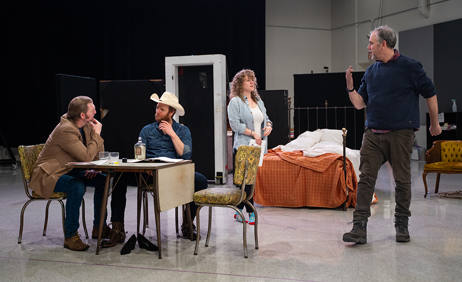 Caroline, Nick, and Cliff receiving direction from director Jeremy Herrin. Nick and Cliff sit at a table, while Caroline and Jeremy stand and talk. Jeremy is a light-skinned man with salt-and-pepper hair. The rehearsal set in the background includes door frames and a bed.