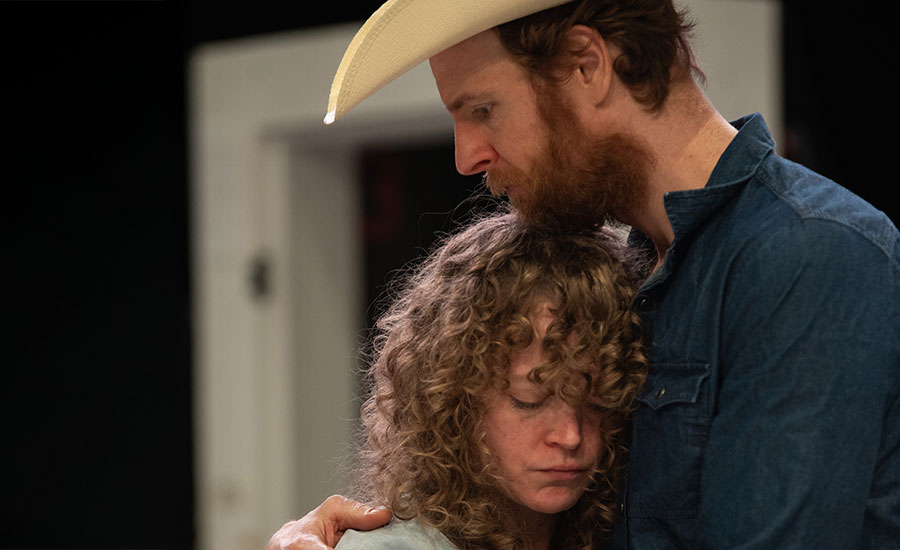 Actors Caroline Neff and Nick Gehlfuss embracing. Caroline is a light-skinned woman with curly red hair. Nick is a light-skinned man with red hair, wearing a jean jacket and cowboy hat.