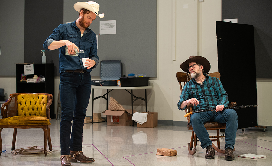 Nick rehearsing with ensemble member Tim Hopper. Tim is a light-skinned man with a salt-and-pepper beard and a cowboy hat. Nick pours alcohol for Tim, who sits in a rocking chair holding a cane.